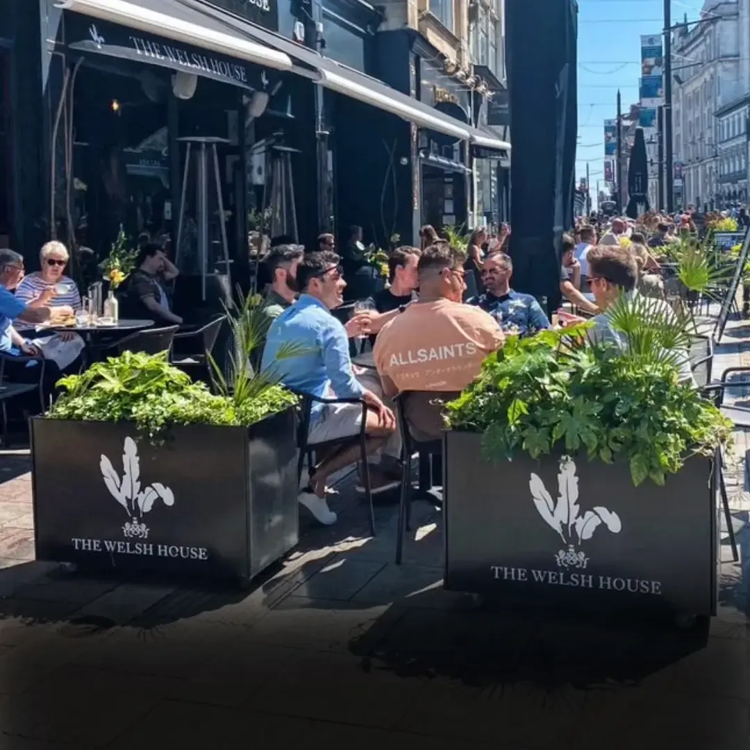 Guests eating and drinking at tables on a busy street terrace, behind planters branded The Welsh House